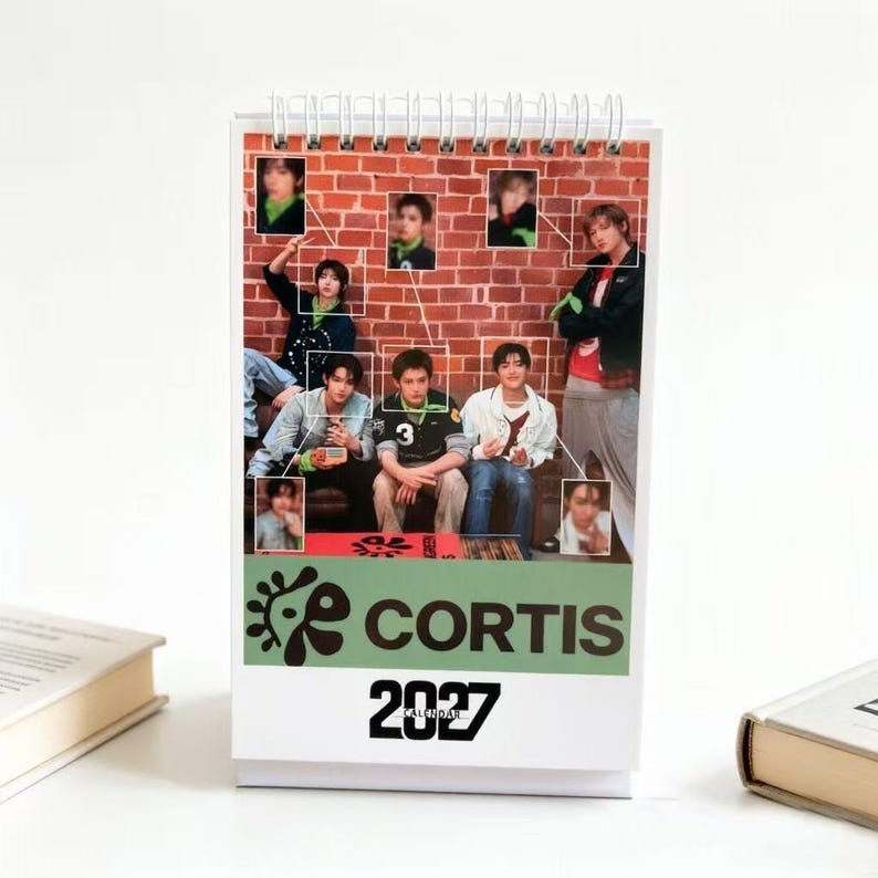 May include: A spiral-bound desk calendar for 2027 featuring a collage of photographs of a group of young people against a red brick wall background. The lower section displays the text CORTIS in bold black letters above the year 2027 in large font.
