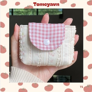 Può includere: Un piccolo portafoglio in tessuto color crema con patta a quadretti rosa e bianchi fissata da un bottone bianco. Il corpo del portafoglio presenta un motivo floreale e a righe. Il marchio "Tomeyavn" è stampato in alto.