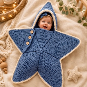 May include: A star-shaped baby swaddle blanket crocheted in dark blue yarn with a cream-colored border. The swaddle features a hood, three wooden buttons on the side flap, and a textured stitch pattern. A small blue bow is attached to the hood.