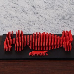 以下が含まれることがあります： 垂直スラットで作られた赤いフォーミュラ1レーシングカーの彫刻で、黒いベースに取り付けられています。ベースにはフォーミュラ1のロゴが見えます。彫刻は濃い木製の表面に展示されています。