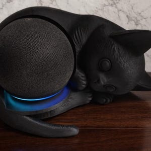 May include: A black cat-shaped speaker holder cradling a black fabric-covered speaker. The cat is in a resting pose, with its head and paws near the speaker. A blue light emanates from the base of the speaker. The holder is on a wooden surface.