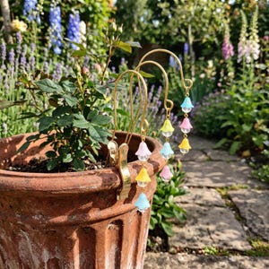 May include: Two gold-toned metal plant stakes with clips, each featuring a vertical chain of three small, pastel-coloured bell-shaped flower charms in blue, pink, and yellow, accented with clear spherical beads hanging from the curved top hooks.