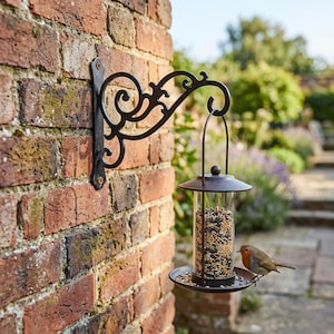 May include: A black metal wall-mounted bracket with an ornate scroll design supports a cylindrical bird feeder. The feeder has a clear plastic tube filled with mixed seeds, a dark metal roof, and a circular base where a small robin is perched.