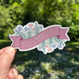 May include: A die-cut sticker featuring a mauve banner with the text MY TUMMY HURTS in white capital letters. The banner is layered over a decorative illustration of light blue and pink flowers with green foliage on a white background.