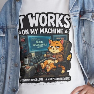 May include: White t-shirt with the text "IT WORKS ON MY MACHINE" and a cartoon cat wearing a hoodie, sitting in a chair in front of a computer. The shirt also has the hashtags #DEVELOPERPROBLEMS and #SLEEPTSFORTHEWEAK.
