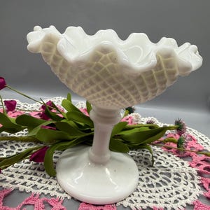 May include: A white, vintage-style compote dish with a ruffled rim and a diamond-patterned body. The dish sits on a pedestal base. The background includes pink and white lace doilies and faux flowers.