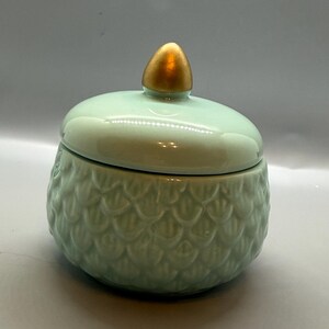 May include: A light mint green ceramic jar with a matching lid featuring a gold-colored, acorn-shaped finial. The body of the jar has a raised, repeating scale-like texture covering the exterior surface, providing a tactile, decorative finish.
