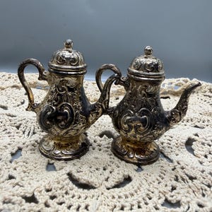 May include: Two antique silver-colored miniature teapots with ornate floral designs. Each teapot has a curved handle, a spout, and a domed lid. The teapots are displayed on a cream-colored, crocheted doily.