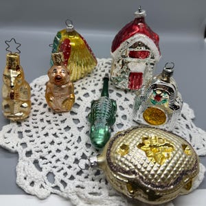 May include: A collection of six vintage glass Christmas ornaments including a gold bear, a monkey, a yellow shell, a red and white house, a green alligator, and a textured gold oval shape with a floral center, all featuring metal hanging loops.