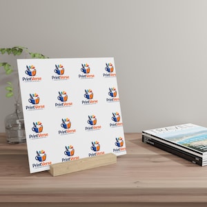 Può includere: Un pannello espositivo quadrato bianco con un motivo ripetuto del logo PrintVerse, che raffigura una tazza da caffè blu e arancione con schizzi di vernice colorati. Il pannello è sostenuto da un supporto in legno chiaro su una superficie piana.