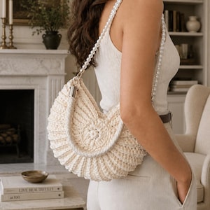 May include: A cream-colored, crescent-shaped crochet handbag featuring a textured, ribbed pattern and a circular center detail. The bag includes a thick, braided handle and a long shoulder strap made of white, spherical faux pearls.
