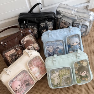 Puede incluir: Una colección de cinco bolsos de hombro rectangulares en negro, plata, marrón, azul claro y crema. Cada bolso cuenta con dos bolsillos frontales con ventanas transparentes que muestran muñecos de peluche, llaveros y tarjetas pequeñas.