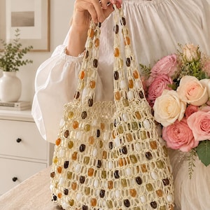 Peut inclure: Un sac fourre-tout en crochet de couleur crème avec un design en filet à mailles ouvertes. Le sac est orné de perles ovales lisses dans des tons blanc, ambre, marron et vert olive, intégrées sur tout le corps et les deux anses supérieures.