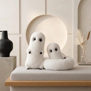 Pode incluir: Duas figuras de pelúcia brancas com grandes olhos negros circulares. Uma está em pé, a outra está sentada em um anel branco e fofo. Os brinquedos estão em um banco de tons neutros com uma moldura de madeira.