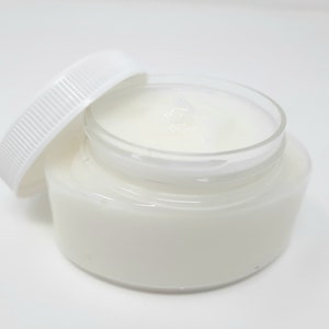 May include: A clear glass jar filled with a white, creamy substance, with the white lid slightly ajar. The jar sits on a white surface, creating a clean and minimalist aesthetic. The product appears to be a skincare cream or balm.
