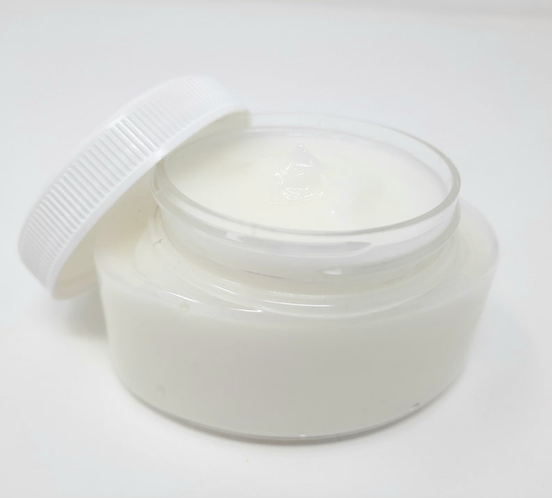 May include: A clear glass jar filled with a white, creamy substance, with the white lid slightly ajar. The jar sits on a white surface, creating a clean and minimalist aesthetic. The product appears to be a skincare cream or balm.