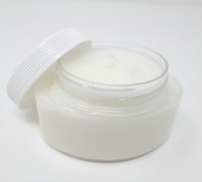 May include: A clear glass jar filled with a white, creamy substance, with the white lid slightly ajar. The jar sits on a white surface, creating a clean and minimalist aesthetic. The product appears to be a skincare cream or balm.