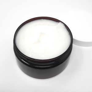 May include: A jar of white cream with a dark brown rim and a white lid. The cream is smooth and appears to be a skincare product. The jar is sitting on a white surface.