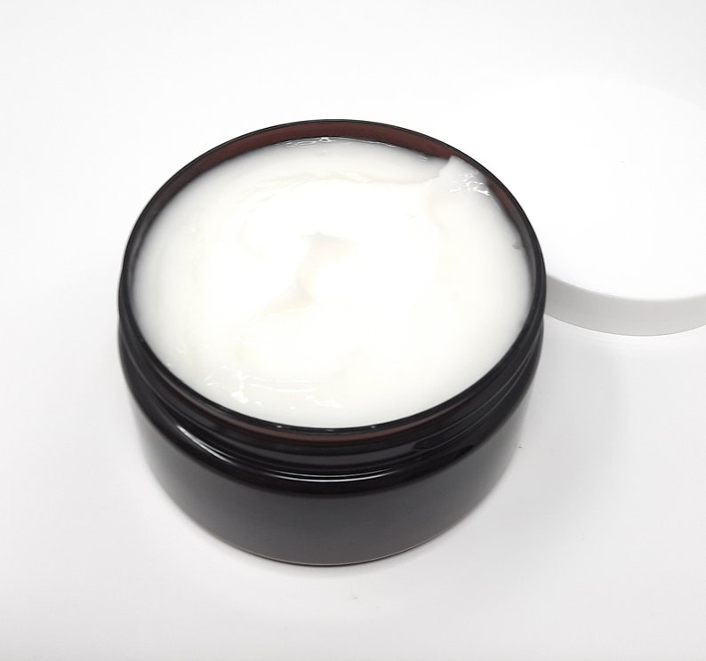 May include: A jar of white cream with a dark brown rim and a white lid. The cream is smooth and appears to be a skincare product. The jar is sitting on a white surface.