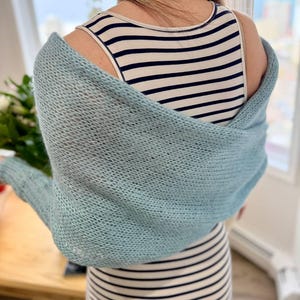Könnte beinhalten: Ein hellblauer, locker gestrickter Bolero mit langen Ärmeln über einem ärmellosen Kleid mit weißen und marineblauen Querstreifen. Der Bolero hat eine weiche, luftige Textur und liegt über den Schultern und dem oberen Rücken.