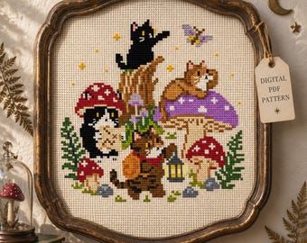 May include: A cross-stitch pattern featuring three cats, mushrooms, and a lantern on beige fabric, framed in a dark wood frame. A tag attached to the frame reads DIGITAL PDF PATTERN. The design includes a black cat, a brown cat, and a tuxedo cat.