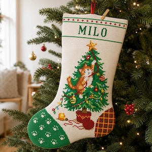 Puede incluir: Un calcetín navideño de punto de cruz en color crema con el nombre MILO en letras verdes en la parte superior. El diseño incluye un árbol de Navidad verde con un gato atigrado trepando, una puntera verde con huellas blancas y un talón a cuadros.