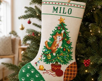以下が含まれることがあります： 上部に緑色の文字でMILOと刺繍されたクリーム色のニードルポイントクリスマスストッキング。デザインには、登っている茶トラ猫が描かれた緑色のクリスマスツリー、白い肉球模様のある緑色のつま先部分、チェック柄のかかとが含まれています。