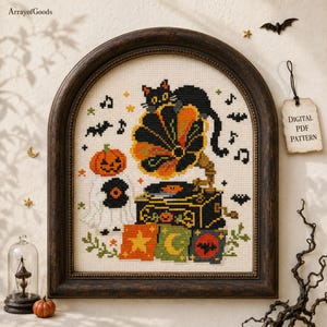 May include: A cross-stitch pattern in a dark wood arched frame featuring a black cat perched on a vintage gramophone, a ghost with a pumpkin head, musical notes, and stars. A tag attached to the frame reads Digital PDF Pattern in black text.