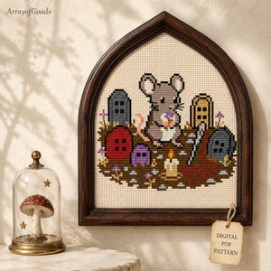 May include: A cross-stitch pattern in a dark wood arched frame featuring a gray mouse holding a flower in a graveyard with tombstones, a candle, and a needle with red thread. A tag attached to the frame reads "DIGITAL PDF PATTERN".