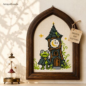May include: A cross-stitch pattern in a dark wood arched frame featuring a green frog wearing a dark cloak standing next to a tall clock tower with a yellow crescent moon clock face. A tag attached to the frame reads DIGITAL PDF PATTERN.