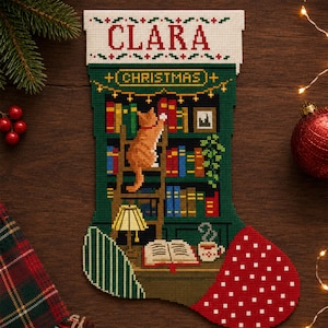 May include: A personalized Christmas stocking featuring a cross-stitch design of a ginger cat on a library ladder reaching for books on shelves. The top cuff is white with the name CLARA in red text, and the toe is red with white polka dots.