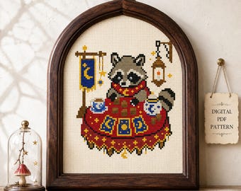 May include: A cross-stitch pattern in a dark wood arched frame featuring a raccoon wearing a red scarf at a table with tarot cards, a blue banner with a crescent moon, and a hanging lantern. A small sign reads DIGITAL PDF PATTERN.
