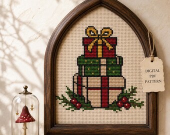 Könnte beinhalten: Ein Kreuzstichmuster in einem dunklen, bogenförmigen Holzrahmen mit einem Stapel aus drei Geschenken in Rot, Grün und Creme mit einer gelben Schleife. Ein Etikett trägt die Aufschrift DIGITAL PDF PATTERN. Eine Glasglocke mit einem Pilz steht daneben.