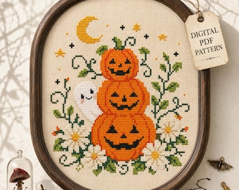 May include: A cross-stitch pattern in an oval wooden frame featuring a stack of three orange jack-o'-lanterns, a small white ghost, a yellow crescent moon, stars, green vines, and white daisies. A tag attached to the frame reads DIGITAL PDF PATTERN.