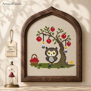 May include: A cross-stitch pattern featuring a brown owl standing under a tree with red apples and hanging lanterns. A basket of apples sits nearby. The design is framed in a dark wood arch. A tag reads DIGITAL PDF PATTERN and ArrayofGoods.