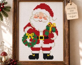 May include: A cross-stitch pattern of Santa Claus wearing a red suit with a white beard, holding a green wreath and a gift. The framed artwork features a tag labeled DIGITAL PDF PATTERN. The design uses a pixelated style on white fabric.