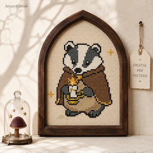 May include: A cross-stitch pattern in a dark wood arched frame featuring a badger wearing a brown cloak and holding a lit candle. The design includes small yellow stars on a cream-colored fabric. A tag reads DIGITAL PDF PATTERN.