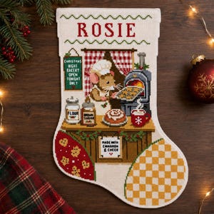 May include: A cream-coloured Christmas stocking featuring a cross-stitch design of a mouse baker in a kitchen. The name ROSIE is stitched at the top. The scene includes a bakery sign, flour, brown sugar, a cinnamon roll, and a red mug.
