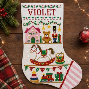 Pode incluir: Uma meia de Natal branca com bordado em ponto cruz e o nome VIOLET no topo. O design inclui uma casa cor-de-rosa, um urso de peluche castanho, um quebra-nozes, um cavalo de baloiço, um tambor, um comboio e bandeirolas coloridas.