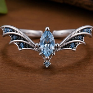 May include: A silver-toned ring featuring a central marquise-cut light blue gemstone. The band is shaped like stylised wings extending outwards, accented with small, dark blue round-cut stones embedded along the wing-like metal structure.