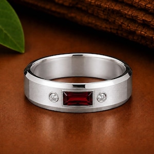 May include: A silver-toned band ring featuring a central rectangular baguette-cut deep red gemstone flanked by two small round clear stones set into the metal surface. The ring has a smooth, polished finish with beveled edges along the band.