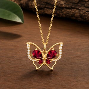 Può includere: Una collana color oro con un ciondolo a forma di farfalla. Il ciondolo è decorato con quattro pietre rosso scuro a forma di pera che formano le ali e piccole pietre chiare che rivestono i bordi esterni delle ali su una catenina sottile.