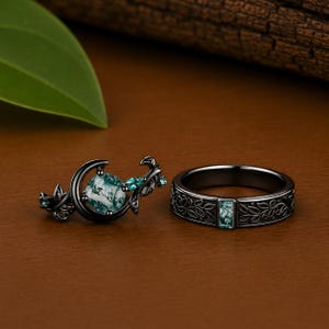 May include: A black metal ring set featuring a crescent moon design with a round moss agate stone and leaf accents, paired with a matching band featuring an engraved vine pattern and a rectangular moss agate inlay set in black metal.