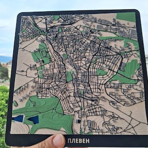 Peut inclure: Une carte en relief 3D carrée de la ville de Pleven, présentant des structures de bâtiments gris en relief, des réseaux routiers noirs et des zones de parcs vertes sur une base beige. La carte est montée sur un cadre noir avec le texte cyrillique ПЛЕВЕН.