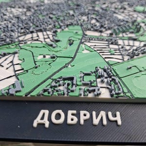 May include: A 3D-printed topographical map of Dobrich, Bulgaria, with green parks, grey buildings, and white roads. The city name, "ДОБРИЧ", is displayed in white letters on a dark blue base.