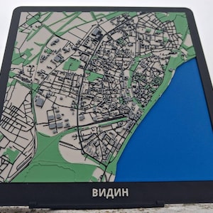 Puede incluir: Un mapa 3D de Vidin, Bulgaria, con marco y soporte negros. El mapa presenta un plano de calles detallado en negro, áreas verdes y una sección azul que representa el río Danubio. La palabra "ВИДИН" está impresa en la parte inferior.
