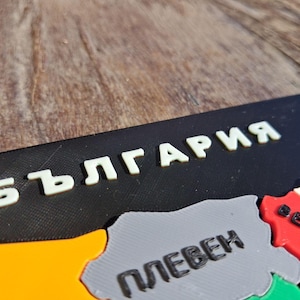 May include: A 3D printed map puzzle of Bulgaria featuring the Cyrillic text БЪЛГАРИЯ in white letters on a black background. The puzzle pieces are colored in yellow, grey, red, and green, with black Cyrillic text labels for city names.