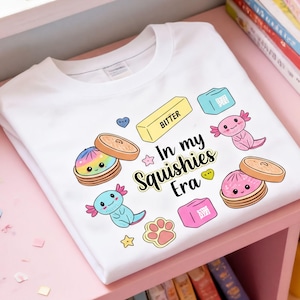 May include: A white t-shirt featuring a colorful graphic print of two axolotls, two bamboo steamers with dumplings, a stick of butter labeled BITTER, and other small icons. The text In my Squishies Era is printed in the center in black.
