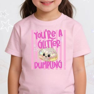May include: A light pink short-sleeved t-shirt featuring a graphic print with vertical pink stripes and the text "YOU'RE A GLITTER DUMPLING" in pink letters. The design includes a central iridescent, sparkling dumpling character with a smiling face.