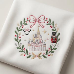 Peut inclure: Un sweat-shirt blanc avec une broderie représentant une couronne de Noël. La couronne comprend un nœud rose, un château, un casse-noisette et une icône d'oreilles de souris. Le motif utilise des fils vert, rose, rouge et doré pour le détail.